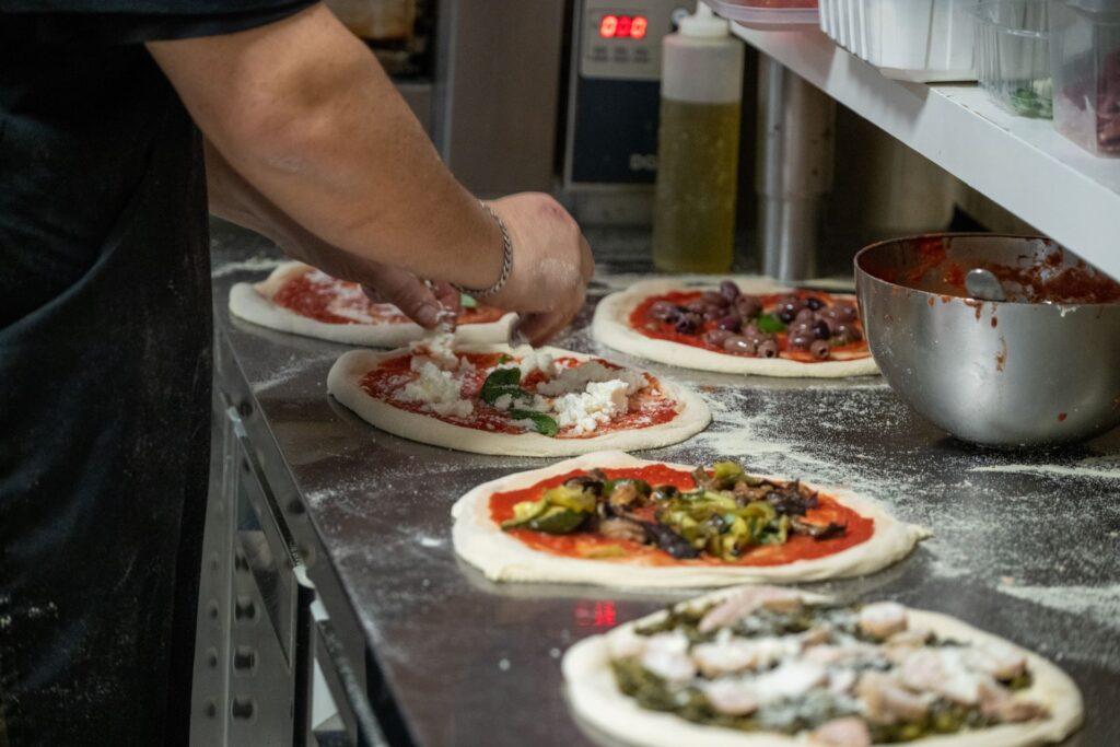 Koch bereitet Pizza an einem Pizzakühltisch mit frischen Zutaten zu.