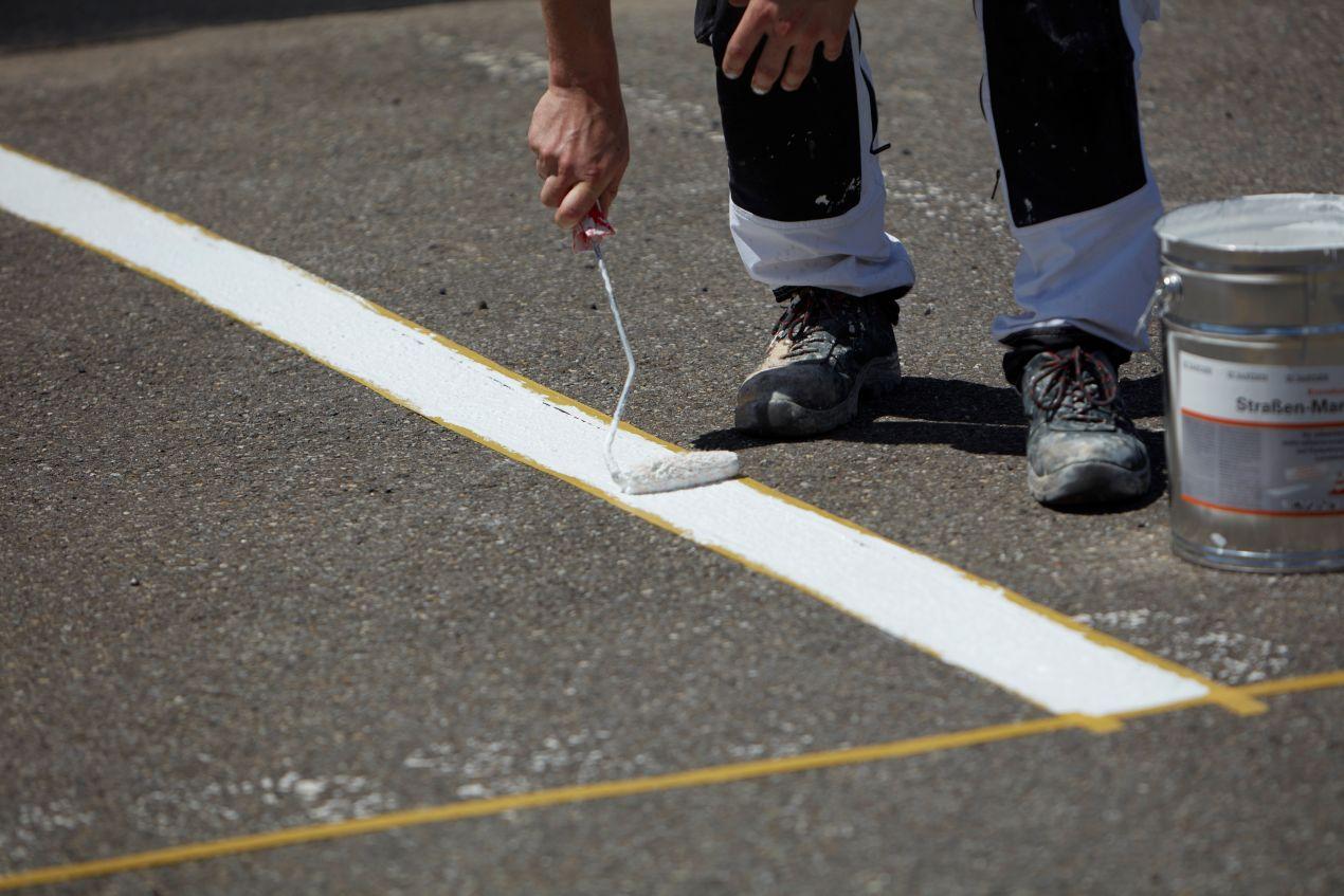 Arbeiter traegt mit Farbrolle weisse Linie auf Asphalt auf – Vorbereitung fuer langlebige Parkplatzmarkierung E-Auto.